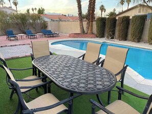 A poolside area with a round table and chairs, a pool, and a view of a house and palm trees. - Trendy and  Tranquil (Las Vegas)