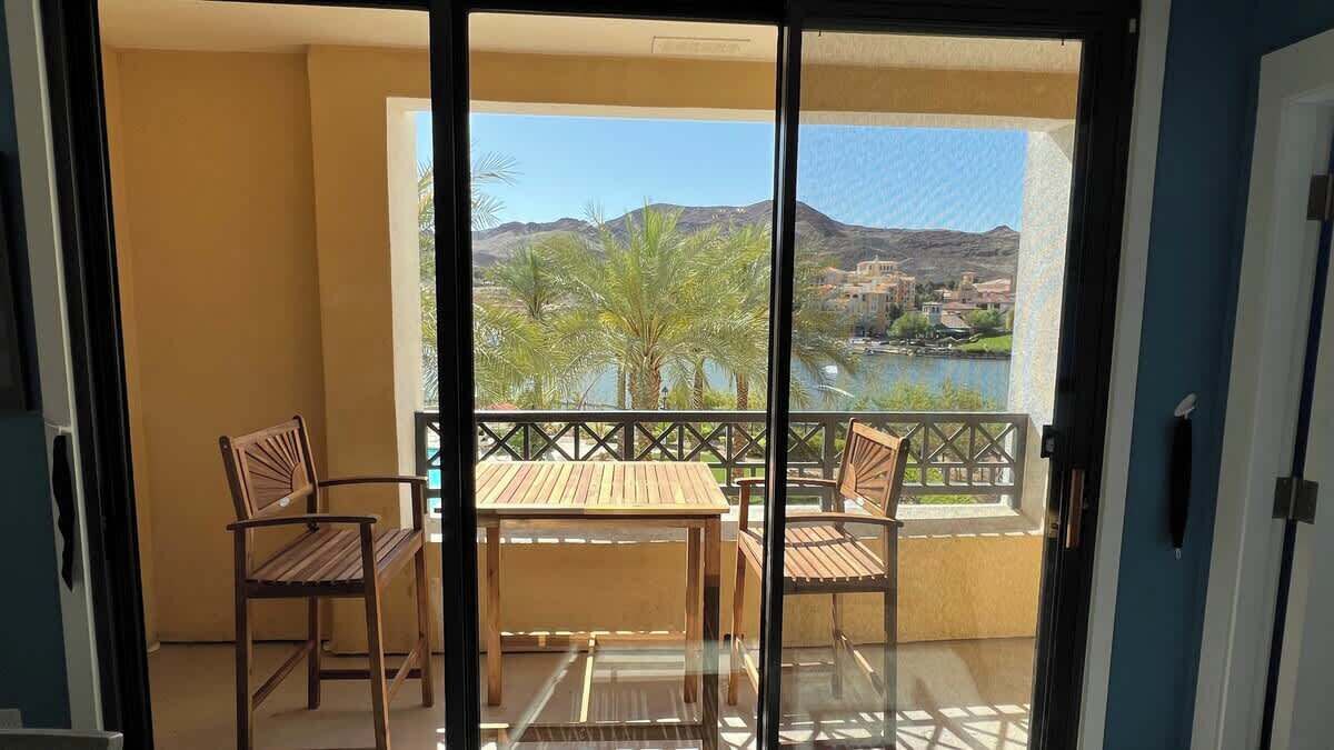 A balcony with a table and chairs, a view of a body of water, and palm trees.