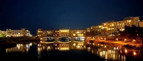 A nighttime view of a waterfront area with illuminated buildings and a bridge reflecting on the water.