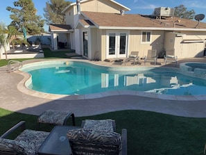 A swimming pool with a curved edge, surrounded by a lawn and a patio area with outdoor furniture. - Spacious Modern Home - 4 miles from strip! (Las Vegas)