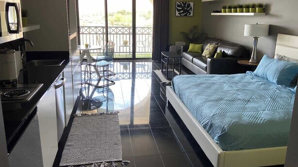A modern hotel room with a balcony, a bed, a sofa, a dining table, and a kitchen area.