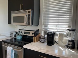 Eine Küche mit Mikrowelle, Toaster, Wasserkocher, Kaffeemaschine, Mixer und einem Fenster mit Jalousien.
