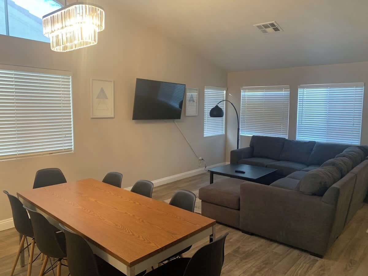 A living room with a wooden dining table, grey sofa, and a TV mounted on the wall.