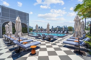 A rooftop pool area with lounge chairs and umbrellas, a checkered floor, and a city skyline in the background.