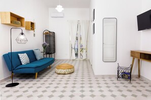 Ein modernes Wohnzimmer mit einem blauen Sofa, einem runden Hocker, einer Stehlampe, einem Fernseher und einem Holzregal.