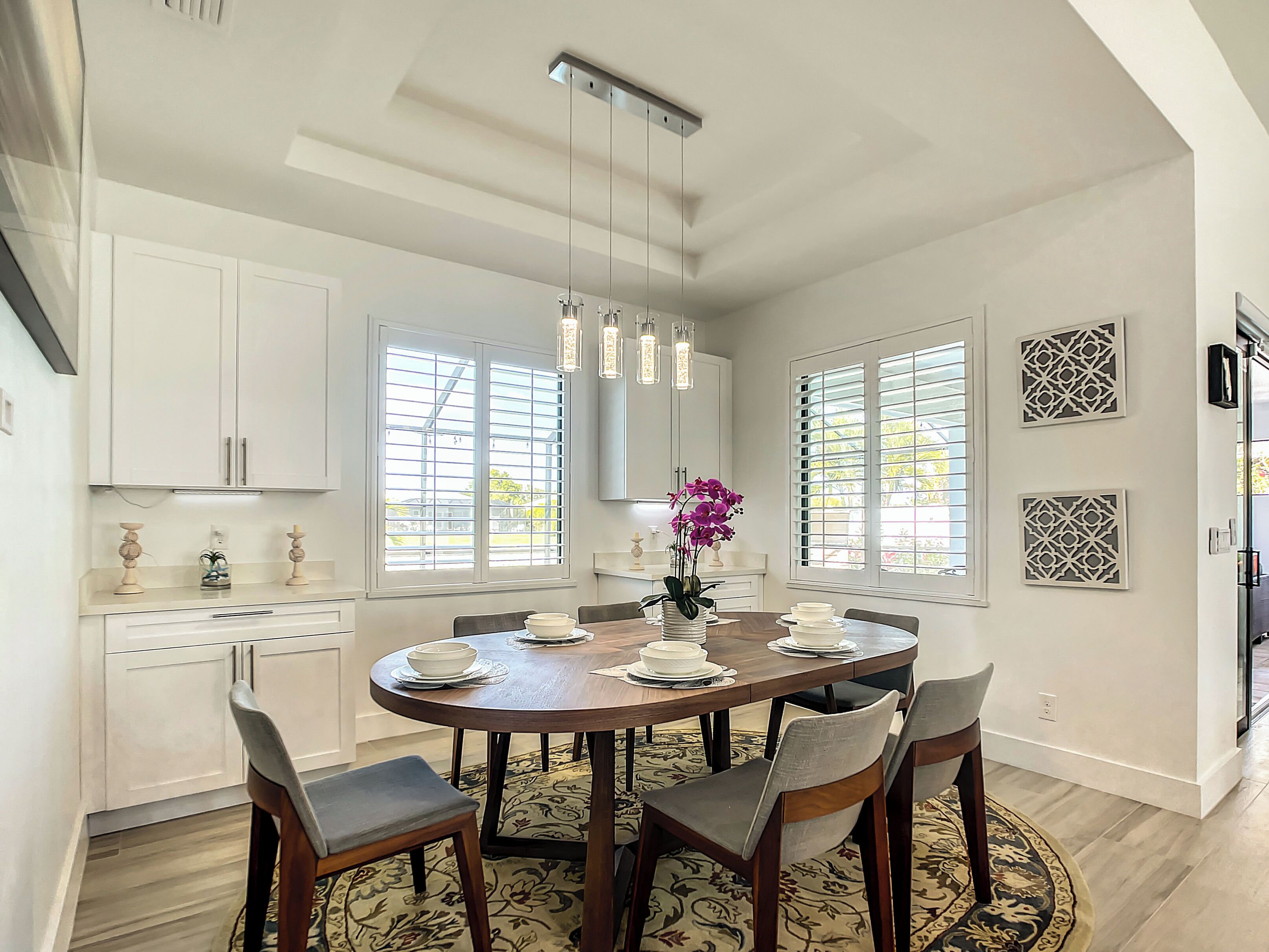 Un comedor con una mesa redonda, sillas y un florero. Hay ventanas con persianas, una lámpara de techo y paneles decorativos en la pared.