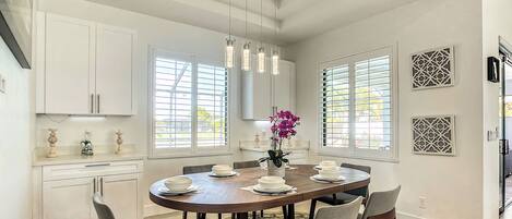 Un comedor con una mesa redonda, sillas y un florero. Hay ventanas con persianas, una lámpara de techo y paneles decorativos en la pared.