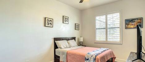 A bedroom with a bed, a ceiling fan, a window with blinds, and a desk with a lamp.