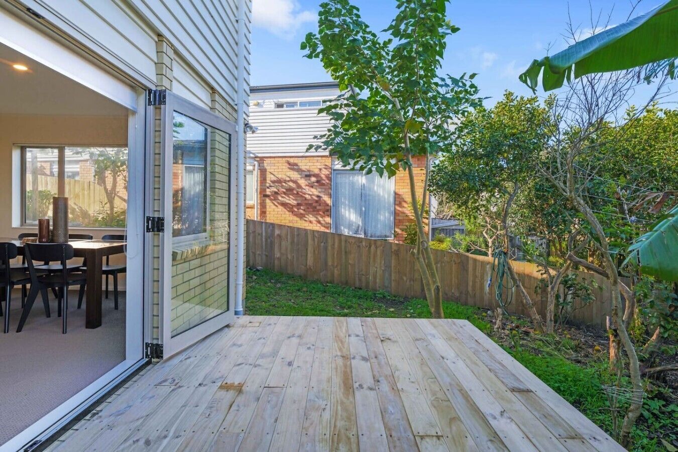 A patio with a wooden deck, a dining area with chairs and a table, and a view of a backyard with a wooden fence and trees.