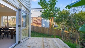 Un patio con terraza de madera, un área de comedor con sillas y una mesa, y vista a un jardín trasero con cerco de madera y árboles.