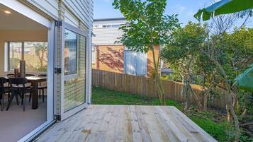 A patio with a wooden deck, a dining area with chairs and a table, and a view of a backyard with a wooden fence and trees.