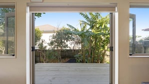 Una puerta corrediza de vidrio da a un patio trasero con terraza de madera, cerca de madera y árboles de plátano.