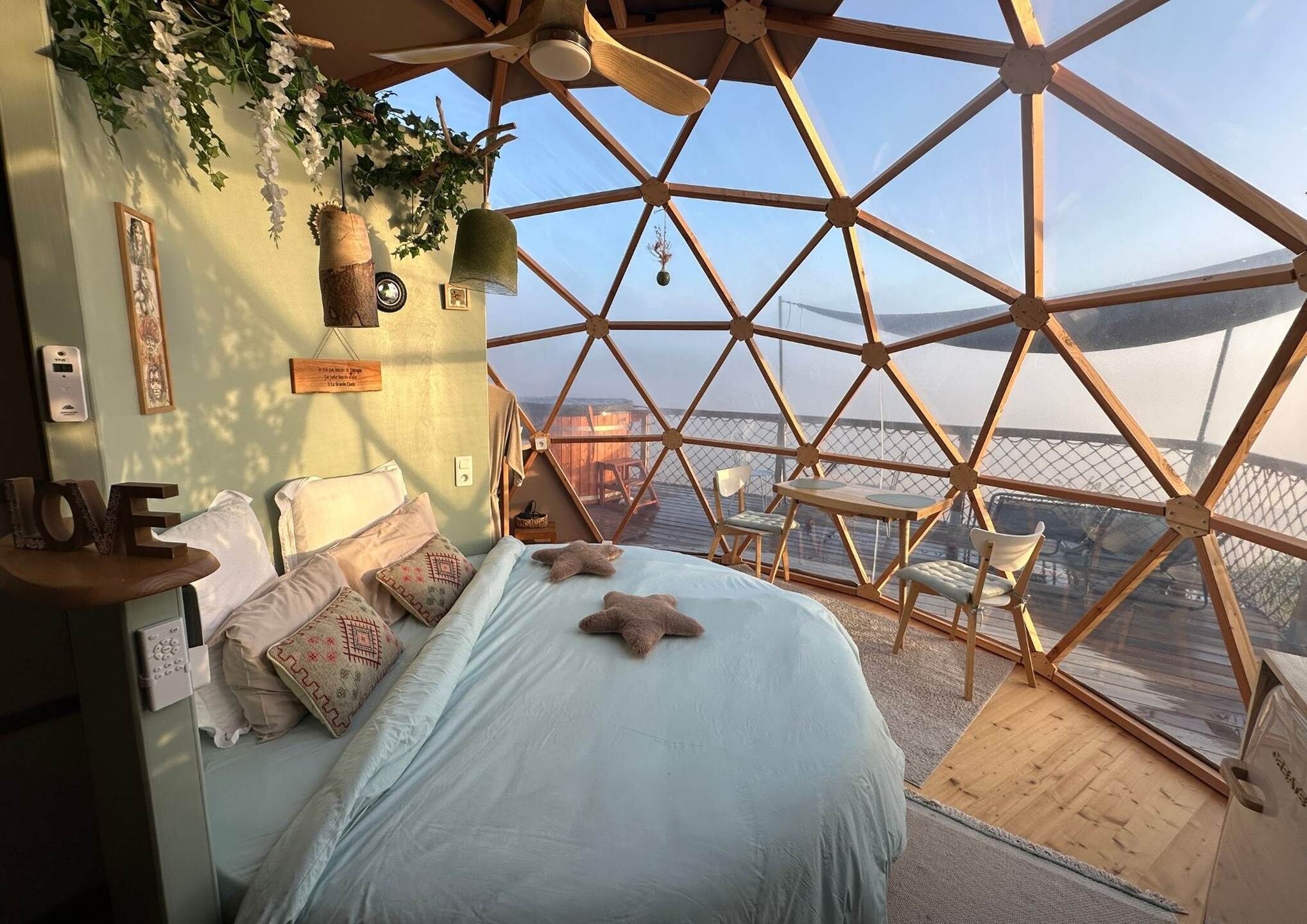 Ein Raum in Form einer Geodätischen Kuppel mit Bett, Kissen, einem kleinen Tisch, Stühlen und Blick nach draußen.