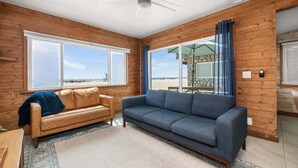 Een kamer met houten inrichting, een groot raam met uitzicht op het strand, een blauwe bank en een bruine leren slaapbank.