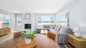 A living room with a fireplace, a sofa, a dining table with a plant, and large windows with a beach view. - Oceanfront Escape With Direct Beach Access (Newport Beach)
