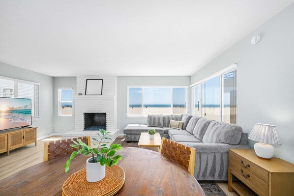 A living room with a fireplace, a sofa, a dining table with a plant, and large windows with a beach view. - Oceanfront Escape With Direct Beach Access (Newport Beach)