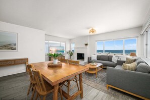 A living room with a fireplace, a dining area with a wooden table and chairs, and large windows with a view of the beach. - Beachfront Duplex Perfect For Large Groups (Newport Beach)