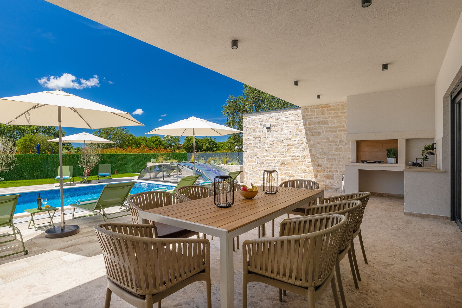 A poolside area with a wooden table, chairs, umbrellas, and a stone wall.