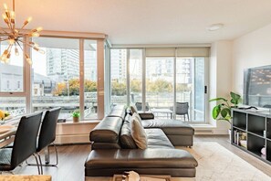 A modern living room with a sofa, a dining table, chairs, a TV, and large windows with a city view. - 5-Min Walk to BC Place, King Bed + Pool & Gym (Vancouver)