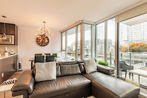 A modern living room with a leather sofa, a dining area, and large windows offering a city view. - 5-Min Walk to BC Place, King Bed + Pool & Gym (Vancouver)