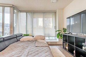A modern bedroom with a large bed, a TV, and a window with a view of the city. - 5-Min Walk to BC Place, King Bed + Pool & Gym (Vancouver)