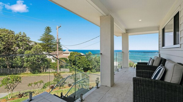 A balcony with a view of the sea, a wicker sofa, and a glass railing. - Glass House Escape at Forresters (Forresters Beach)