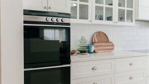 A kitchen with white cabinetry, a black oven, and a countertop with a wooden cutting board and ceramic containers. - Glass House Escape at Forresters (Forresters Beach)