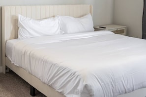 Una cama con ropa de cama blanca, una mesita de noche y un velador.