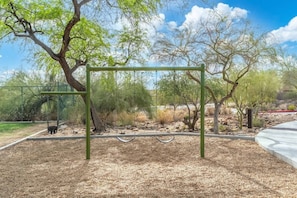 Un parque infantil con columpios y un área cercada, rodeado de árboles y un cielo despejado.