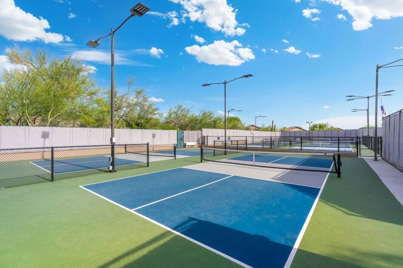 An outdoor tennis court with multiple courts, surrounded by a fence, and equipped with overhead lighting.