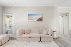 A living room with a beige sofa, a white rug, a painting of a beach, and a white cat.