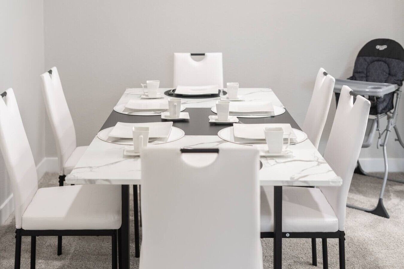 Mesa de jantar posta para seis com cadeiras brancas, um jogo americano preto e pratos e copos brancos.