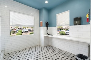 A bathroom with white tiled walls, a patterned floor, a window with blinds, a shelf with a black object, and a fire extinguisher.