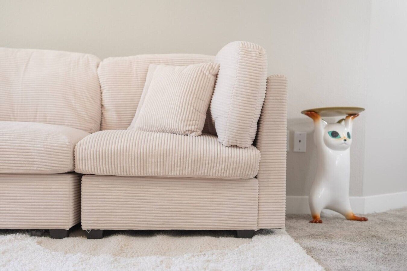 Een beige bank met een witte kussen, een witte katbeeldje, en een witte vloerkleed.