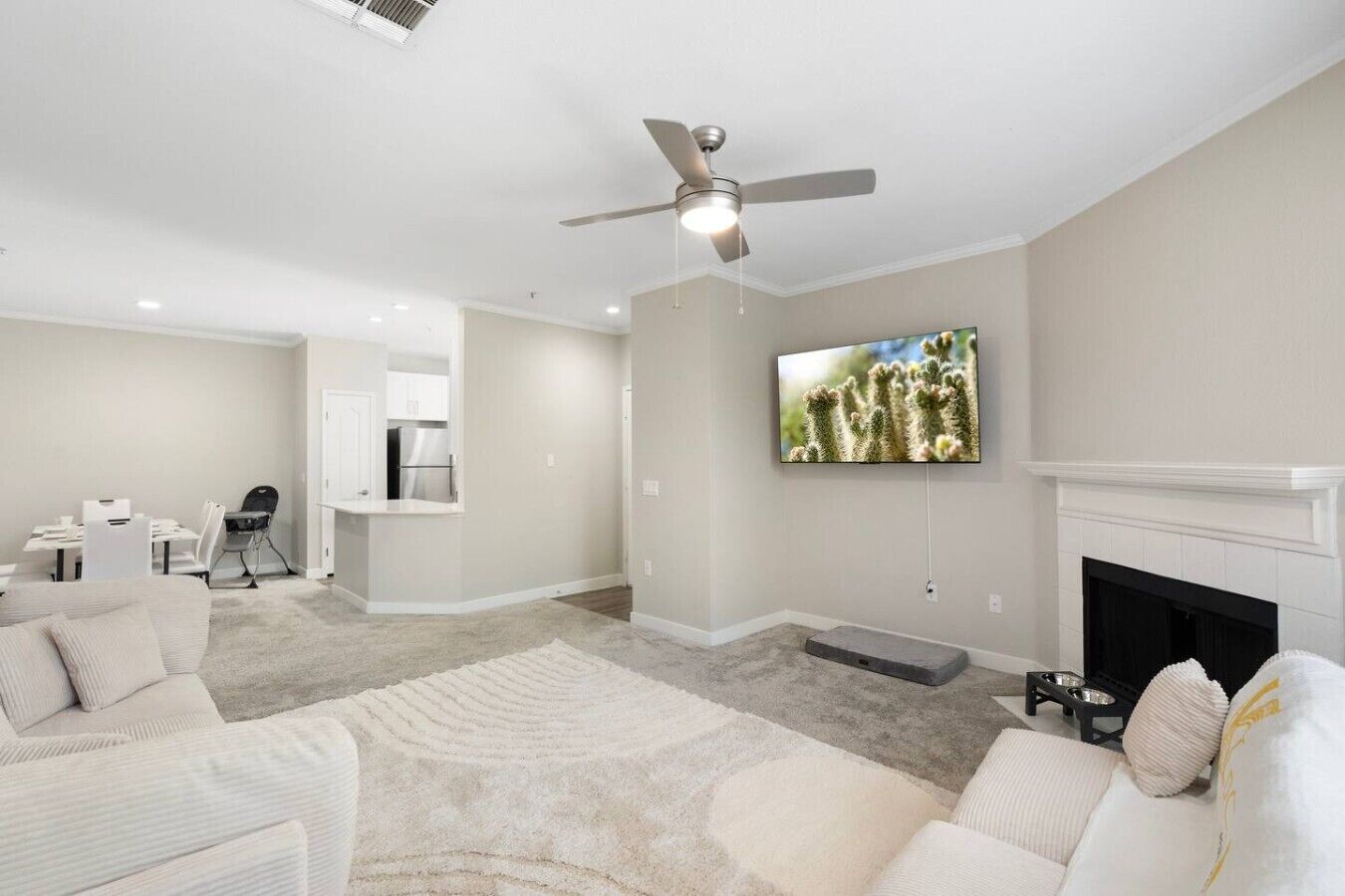 Una sala de estar moderna con ventilador de techo, televisión de pantalla plana, chimenea y una zona de cocina con armarios blancos.