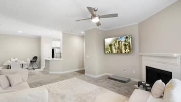 Una sala de estar moderna con ventilador de techo, televisión de pantalla plana, chimenea y una zona de cocina con armarios blancos.