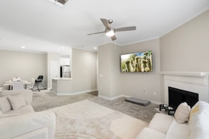 A modern living room with a ceiling fan, a flat-screen TV, a fireplace, and a kitchen area with white cabinets.