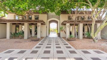 A courtyard with a central pathway leading to an arched entrance, surrounded by trees and potted plants.