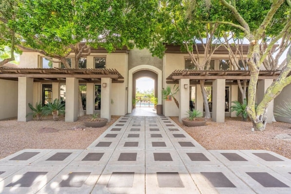 A courtyard with a central pathway leading to an arched entrance, surrounded by trees and potted plants. - Just A Little Comfort (Scottsdale)