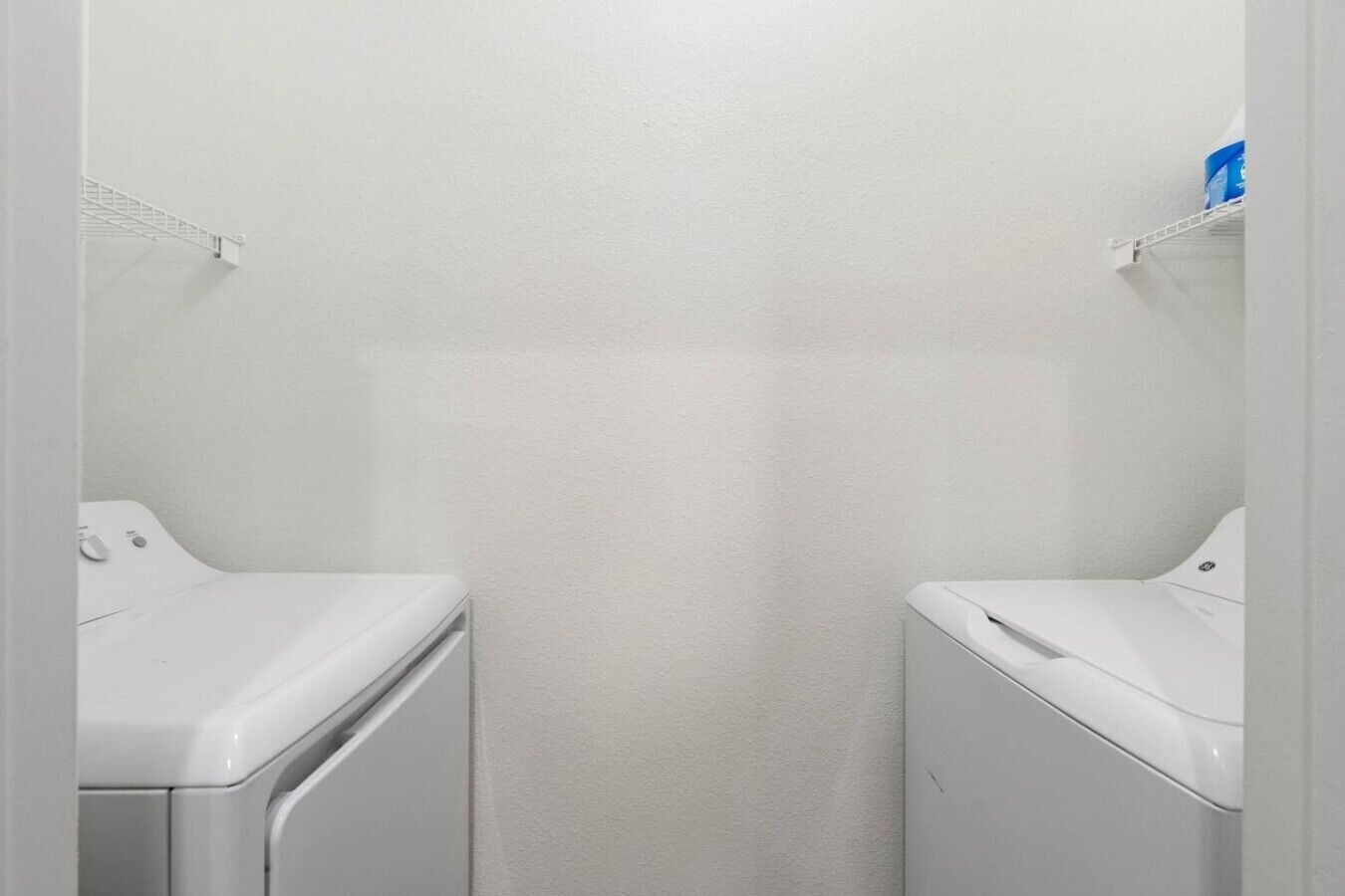 Un lavadero con dos lavadoras blancas, una pared blanca y un estante blanco con un recipiente azul.