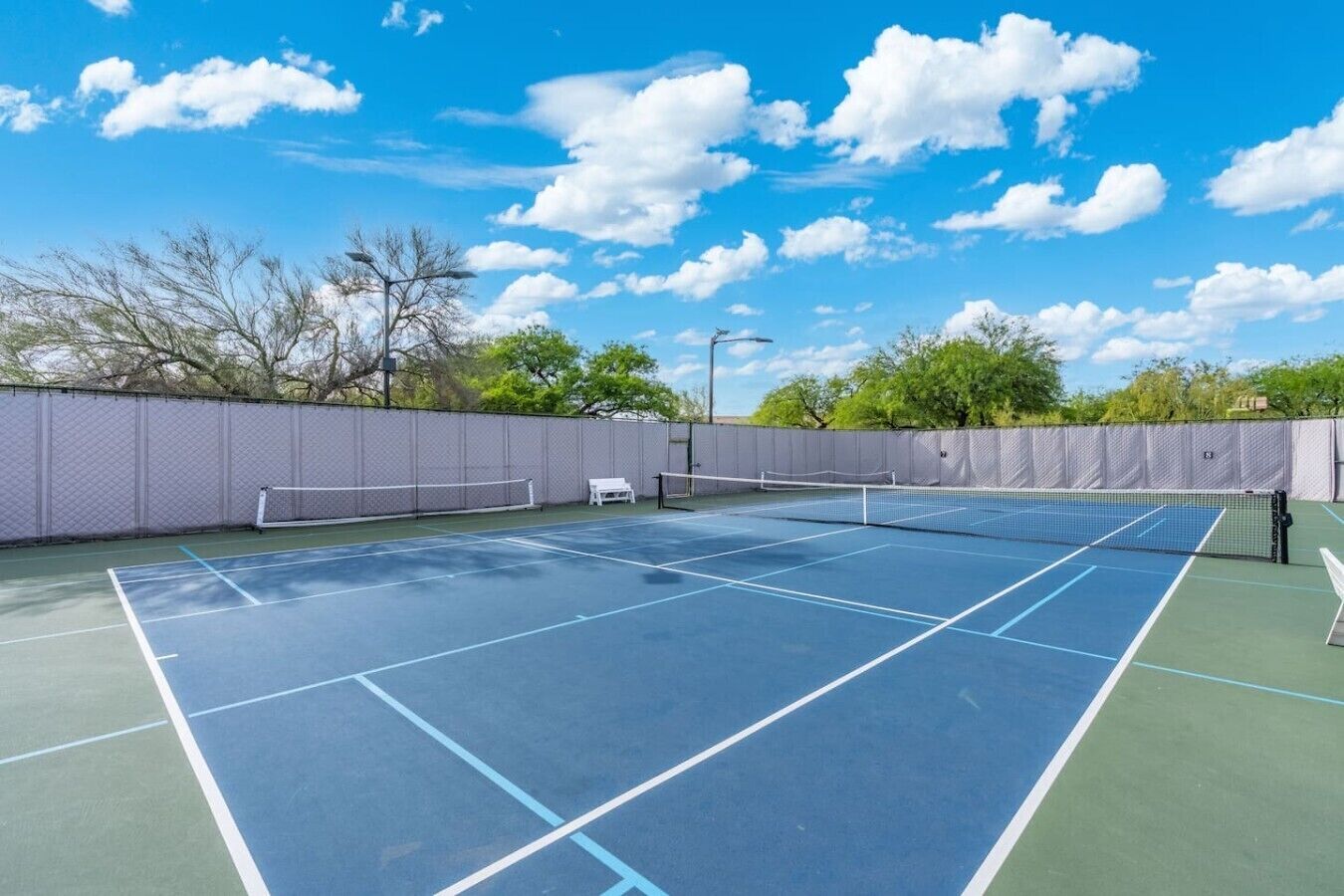 A tennis court with a net, surrounded by a fence, under a clear sky.