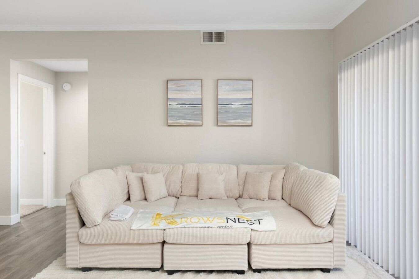 Una sala de estar con un sofá color beige, dos cuadros en la pared y una puerta blanca.