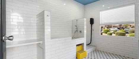 Une salle de bain aux murs recouverts de carreaux blancs, avec une douche, une fenêtre donnant sur un beau paysage et un ventilateur de plafond.