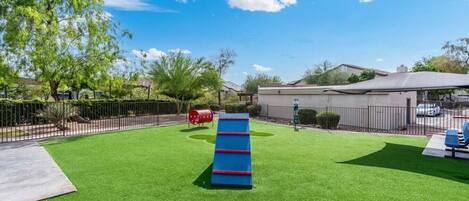 A playground with artificial grass, a blue climbing structure, and a red barrel.