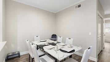 A dining area with a table set for six, white chairs, a black chair, a white ceiling, and a door in the background.