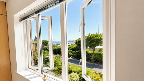 Ein Fenster mit weißen Rahmen, Blick auf eine Straße, Grünflächen und ein weiter Baukörper in der Ferne.