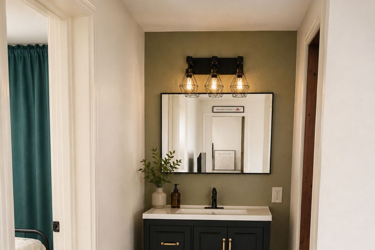 A narrow hallway with a dark wooden vanity, a mirror with three lights, a small plant, and a checkered rug.
