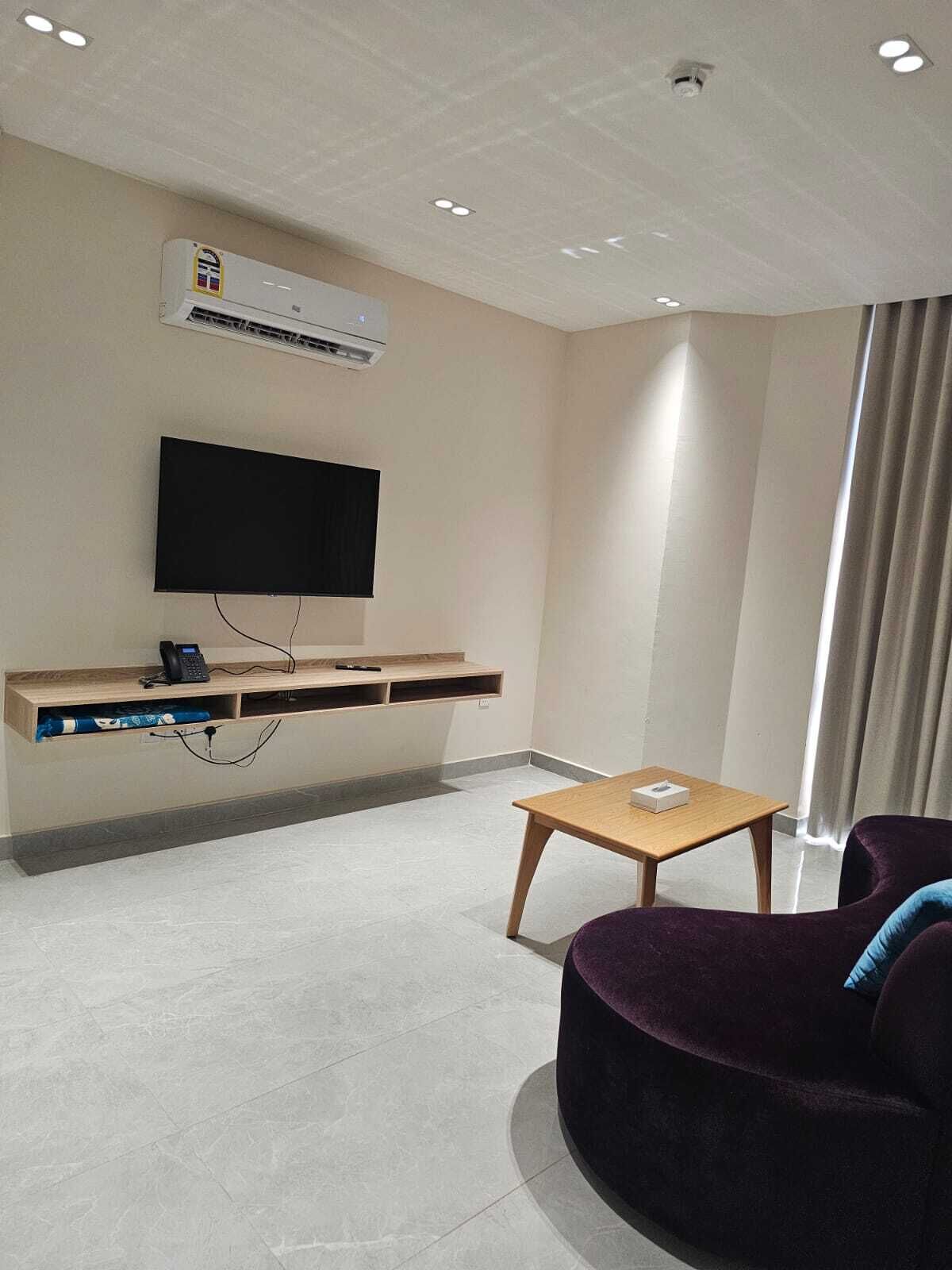 Une chambre d’hôtel équipée d’un téléviseur fixé au mur, d’une table en bois, d’un canapé violet et d’une fenêtre avec des rideaux.