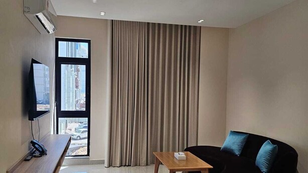 A modern living room with a flat-screen TV, a wooden table, a black sofa with blue cushions, and a large window with curtains.