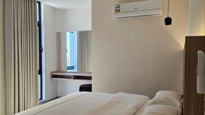 A hotel room with a large bed, an air conditioning unit, a desk with a chair, and a window with curtains.